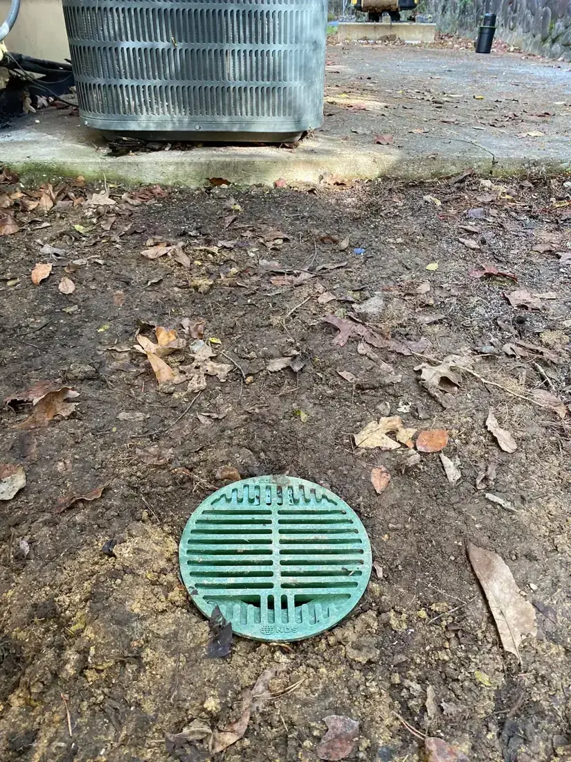Green catch basin grate set in the soil beside an air conditioner pad