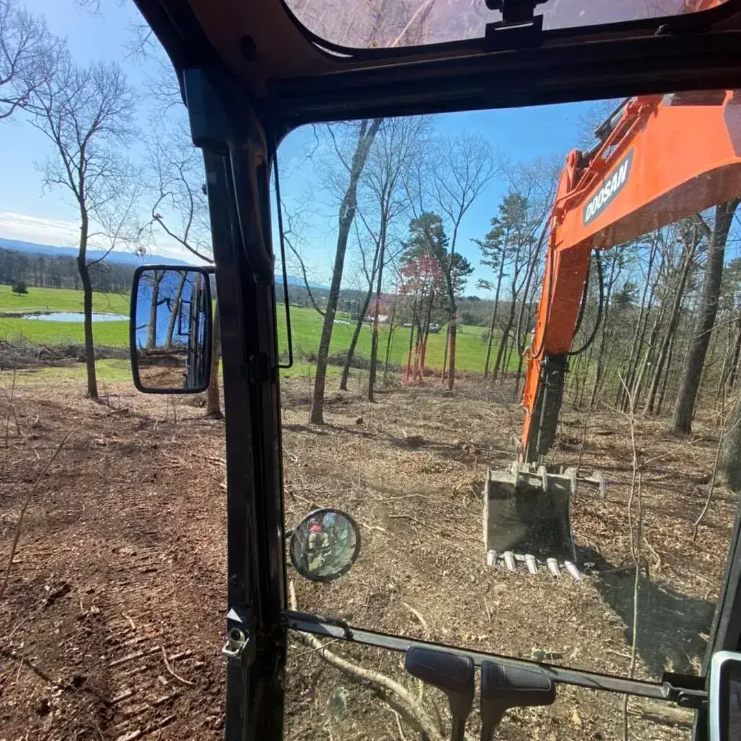 View from an excavator cab over a cleared wooded hillside with pasture and a pond in the distance
