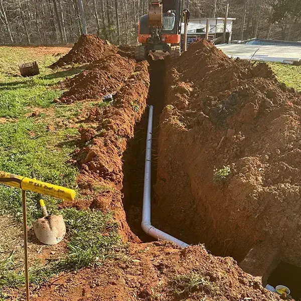 White PVC pipe running down a trench toward a septic tank excavation with an excavator behind