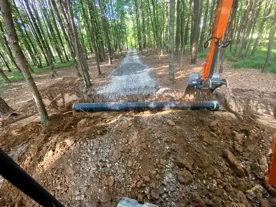 Corrugated drain pipe laid across a graveled path through a wooded lot with an excavator at work