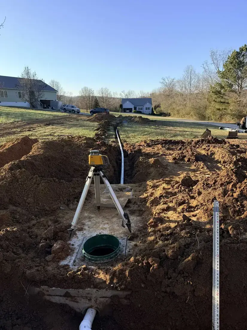 Open septic tank lid with a laser level on a tripod and a pipe running down a long trench to the drain field