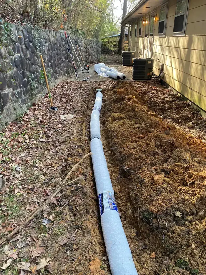 Wrapped drain pipe laid in a trench between a fieldstone retaining wall and the side of a house